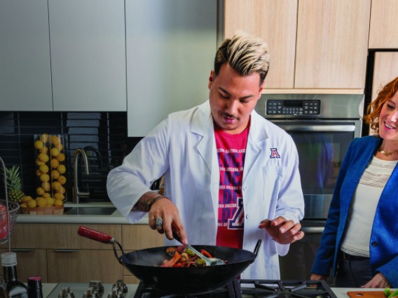 Man wearing white lab coat and woman wearing blue sport coat using a wok to cook in a kitchen