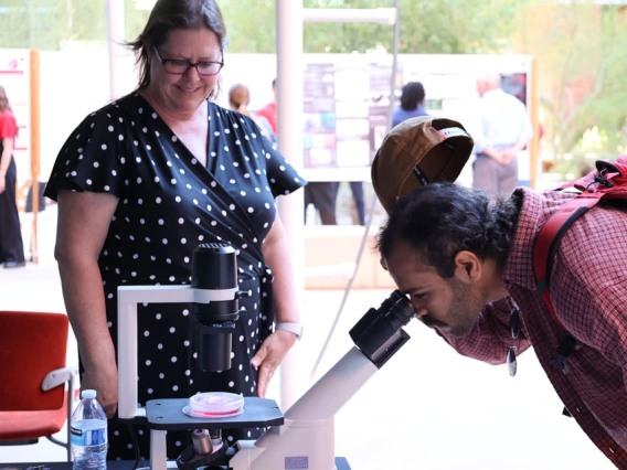 Person looks into a microscope while a researcher smiles and looks on