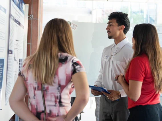 Three people discussing a presentation at a conference poster session. One person, wearing a name badge, is explaining the content on the poster to two attentive listeners.