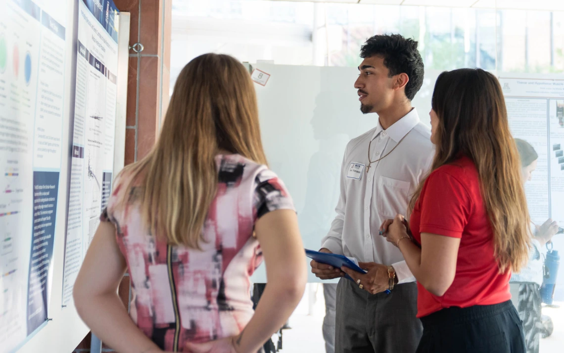 Three people discussing a presentation at a conference poster session. One person, wearing a name badge, is explaining the content on the poster to two attentive listeners.