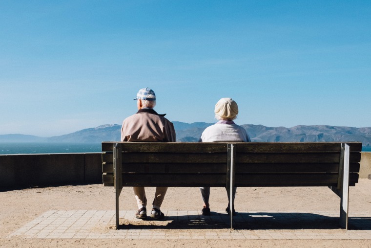 Two elderly people sitting on a bench looking over the horizon with mountains in the distance